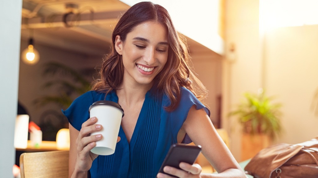 smiling woman looking at her phone and drinking a coffee